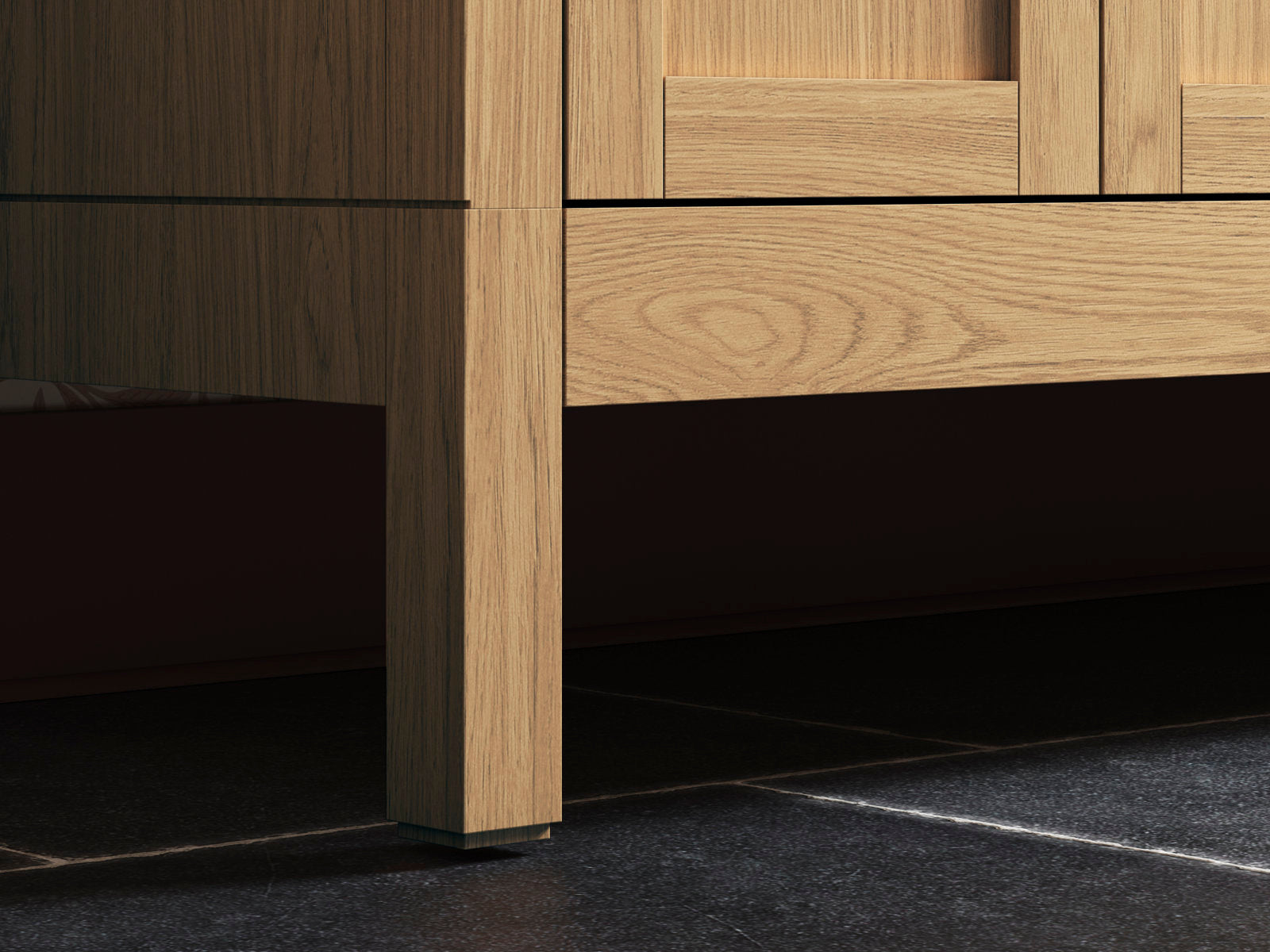 Close-up of the lower corner of a modern solid wood bathroom vanity in a natural wood finish, showing a square leg and drawer apron with visible wood grain above a dark stone tile floor.