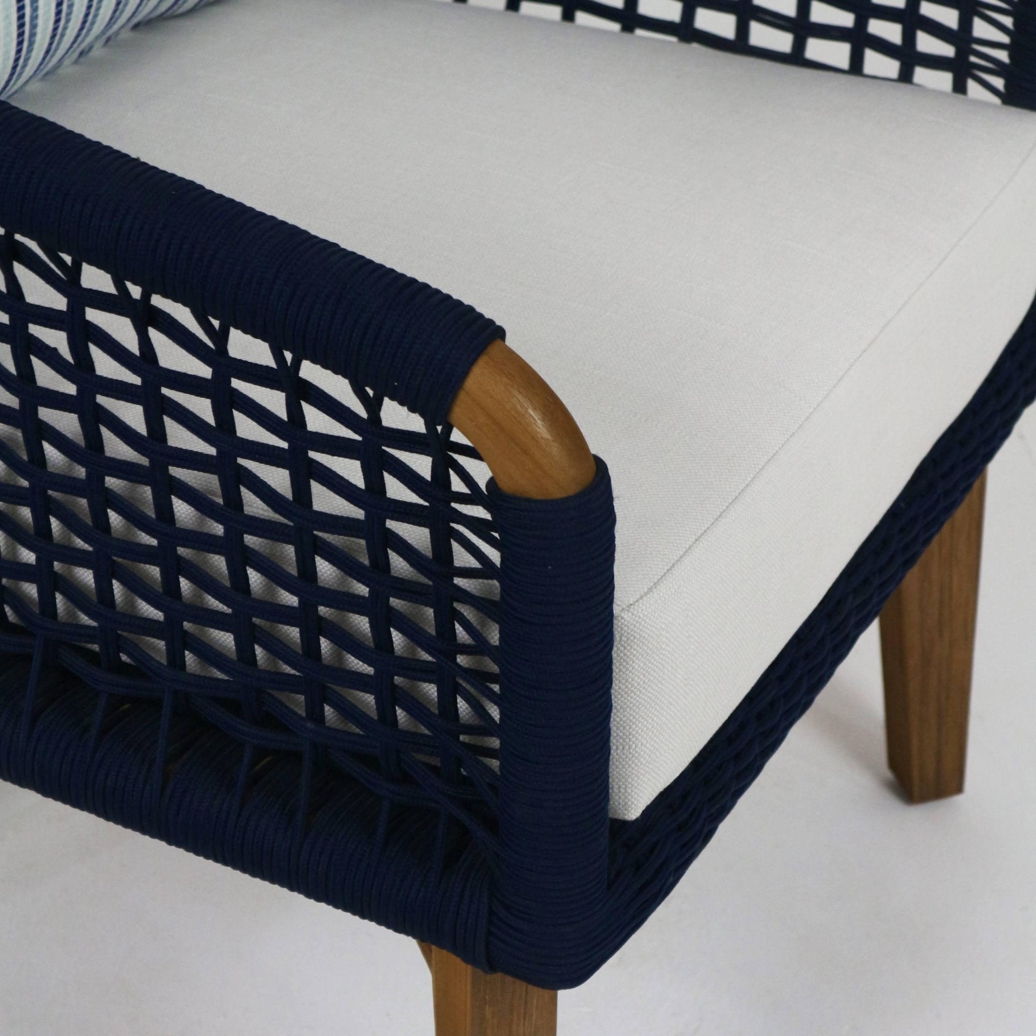 Close-up of an outdoor lounge chair corner featuring navy rope woven back and arm, white fabric seat cushion, and natural wood legs.