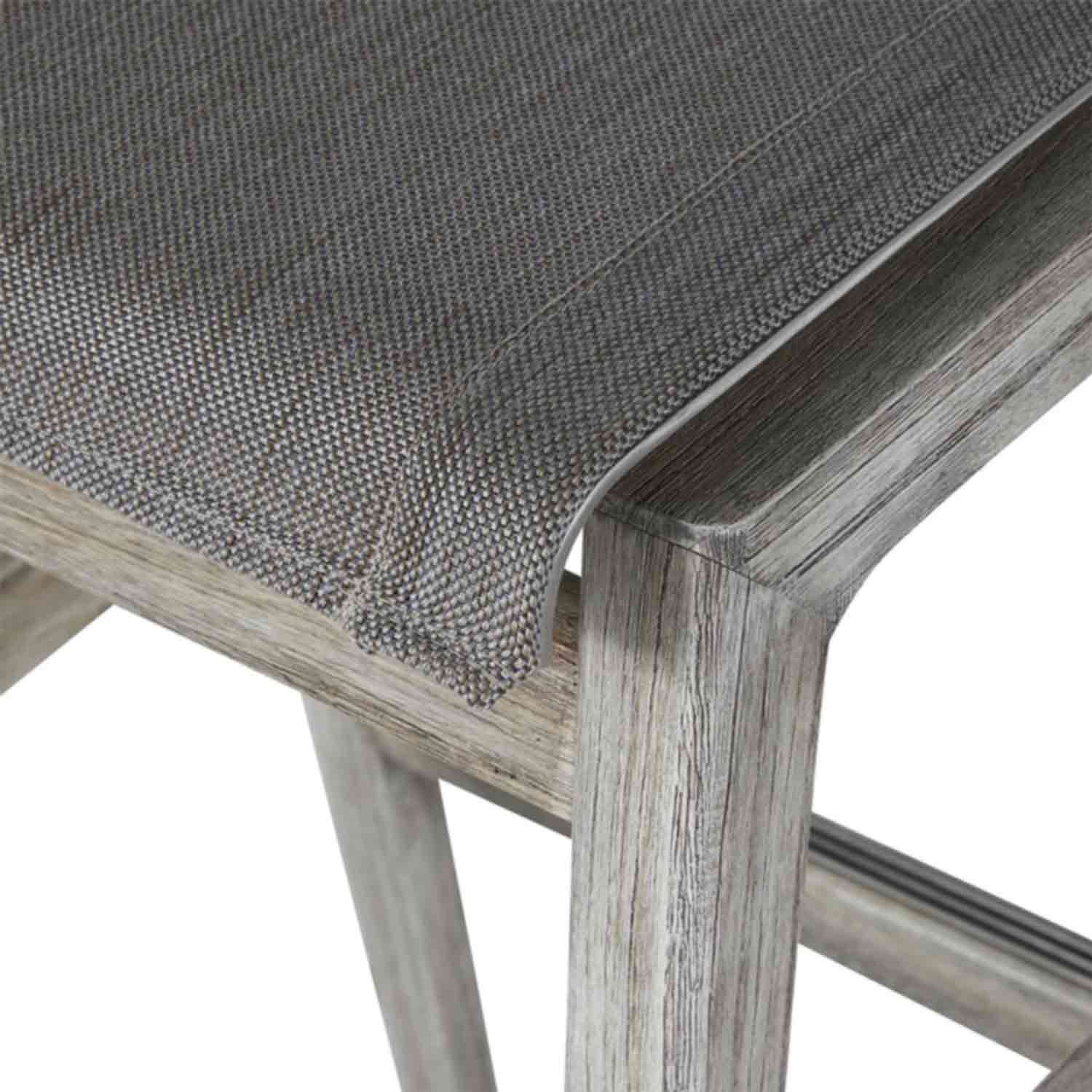 Close-up of a backless outdoor bar stool corner showing gray woven canvas seat stretched over a natural teak frame with visible weathered wood grain and a lower footrest.