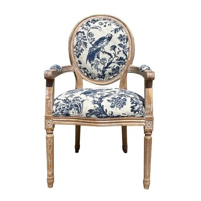 French-style dining armchair with carved teak frame in a natural distressed finish and round medallion back, upholstered in cream linen with a blue toile bird and floral pattern, padded seat and upholstered armrests.