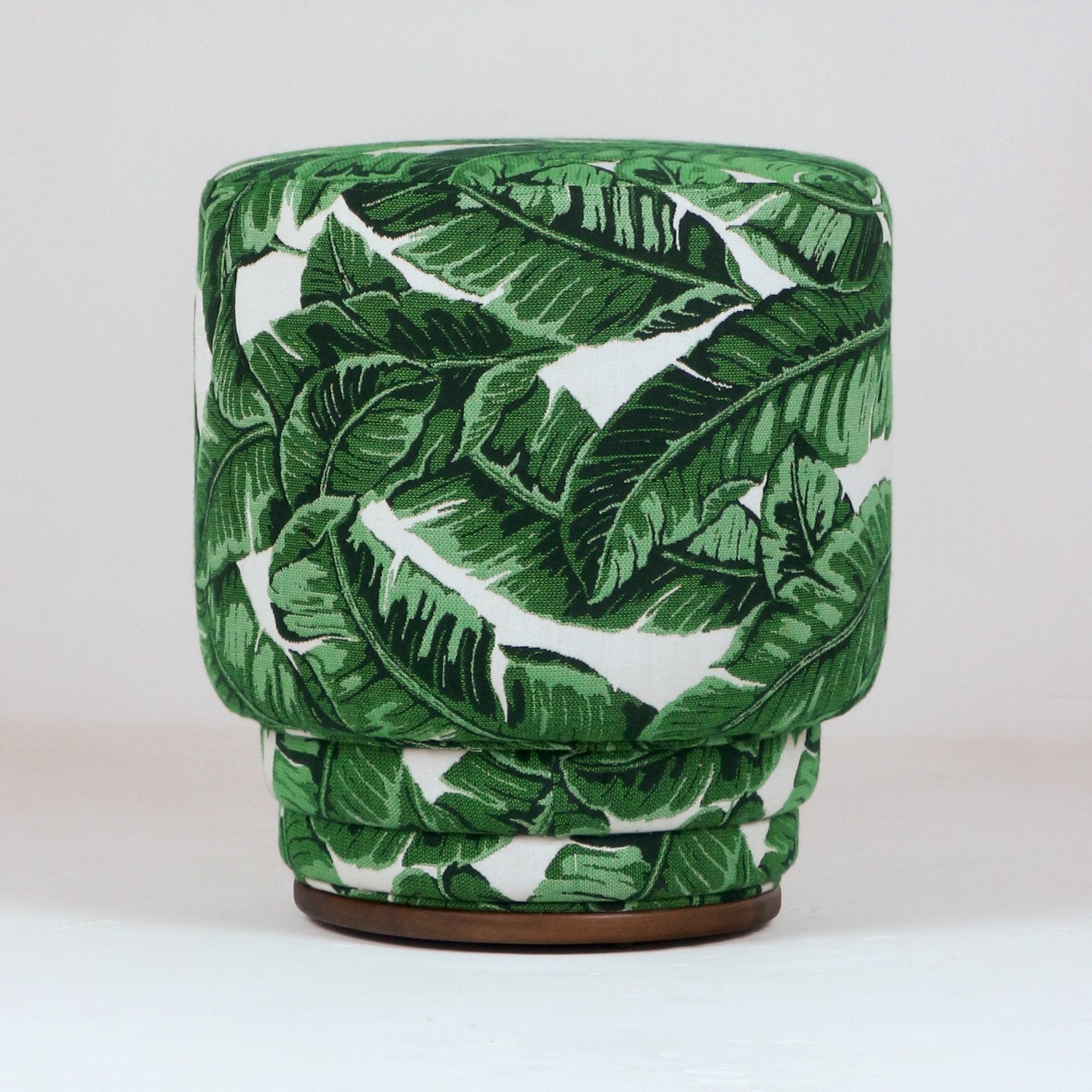 Round modern ottoman upholstered in green tropical leaf fabric with a cushioned top, layered skirted sides and a low wooden circular base.