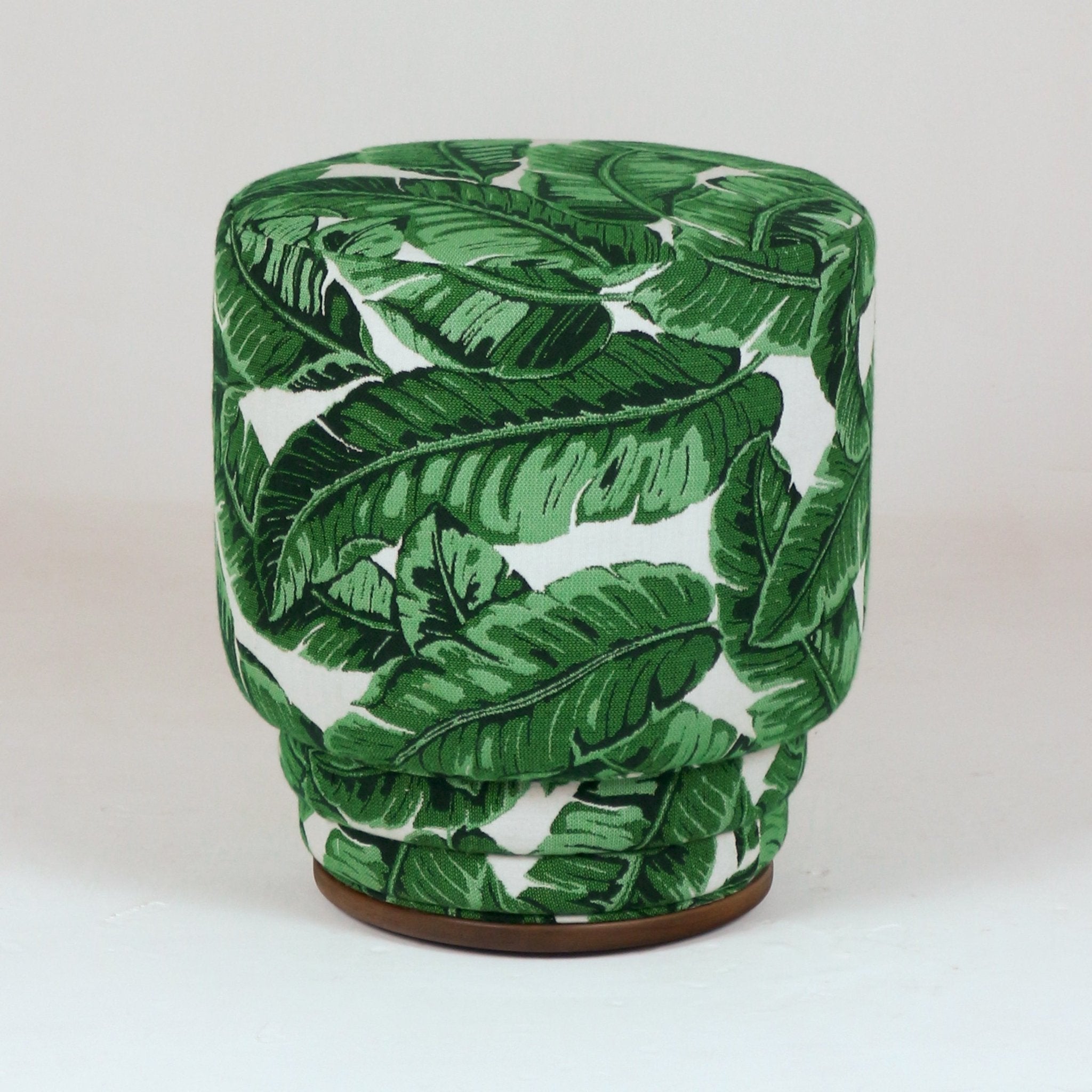 Round modern outdoor upholstered ottoman in green tropical leaf fabric on a white ground, with a cushioned top, pleated sides and a low wooden base.