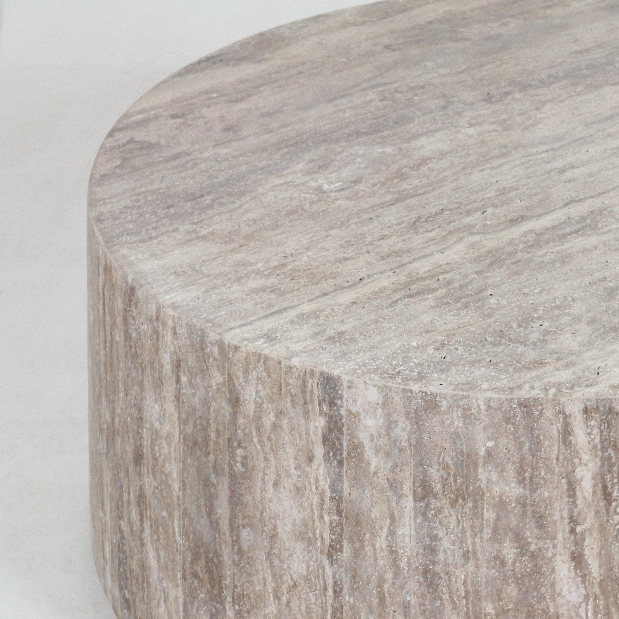 Close-up of a round natural beige travertine coffee table showing the textured stone top and vertically striated side, highlighting the material's veining and matte finish in a modern contemporary style.