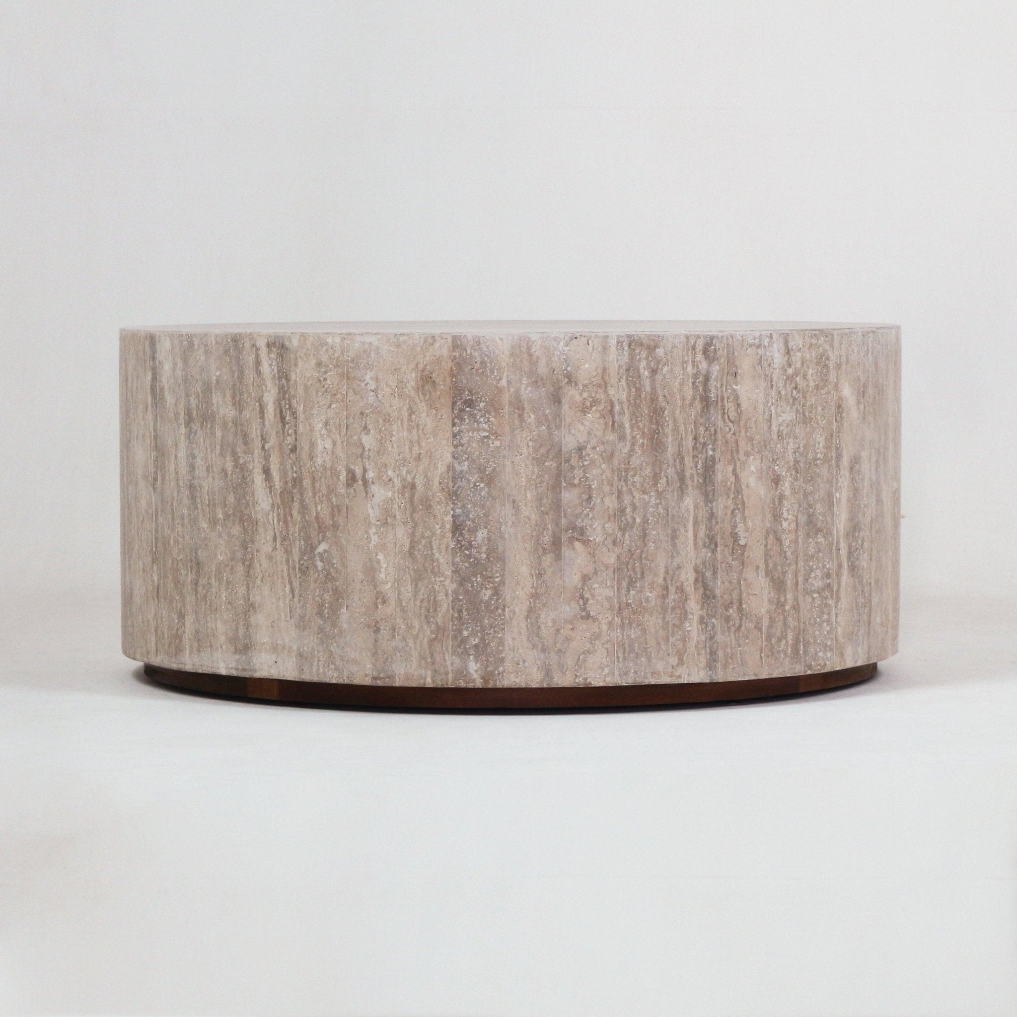 Low round travertine coffee table in natural beige with vertical stone veining and a slim dark wood plinth base, photographed on a white background.