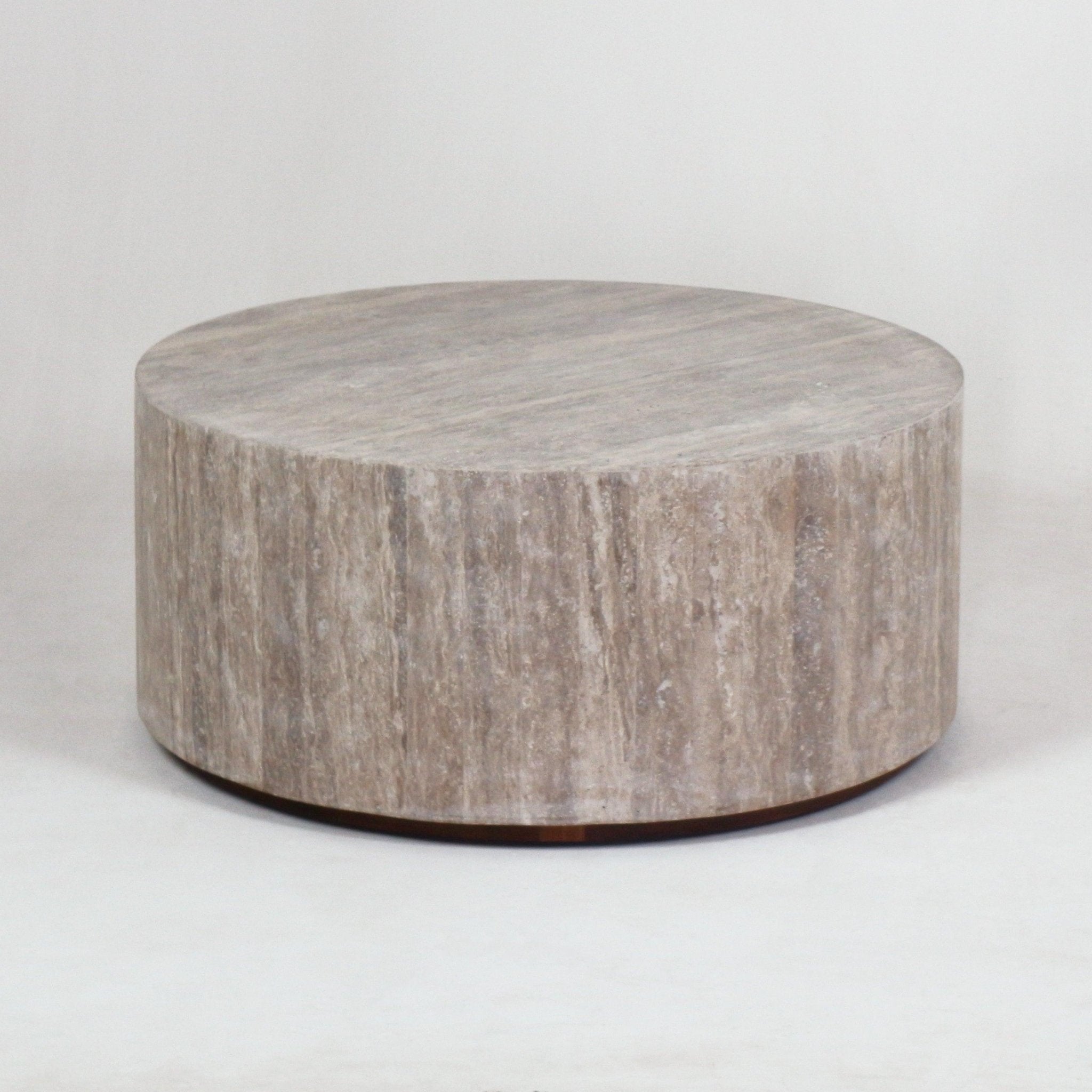 Low round travertine coffee table in natural beige stone with a smooth cylindrical base and subtle dark metal plinth, shown against a neutral background in a modern minimalist style.