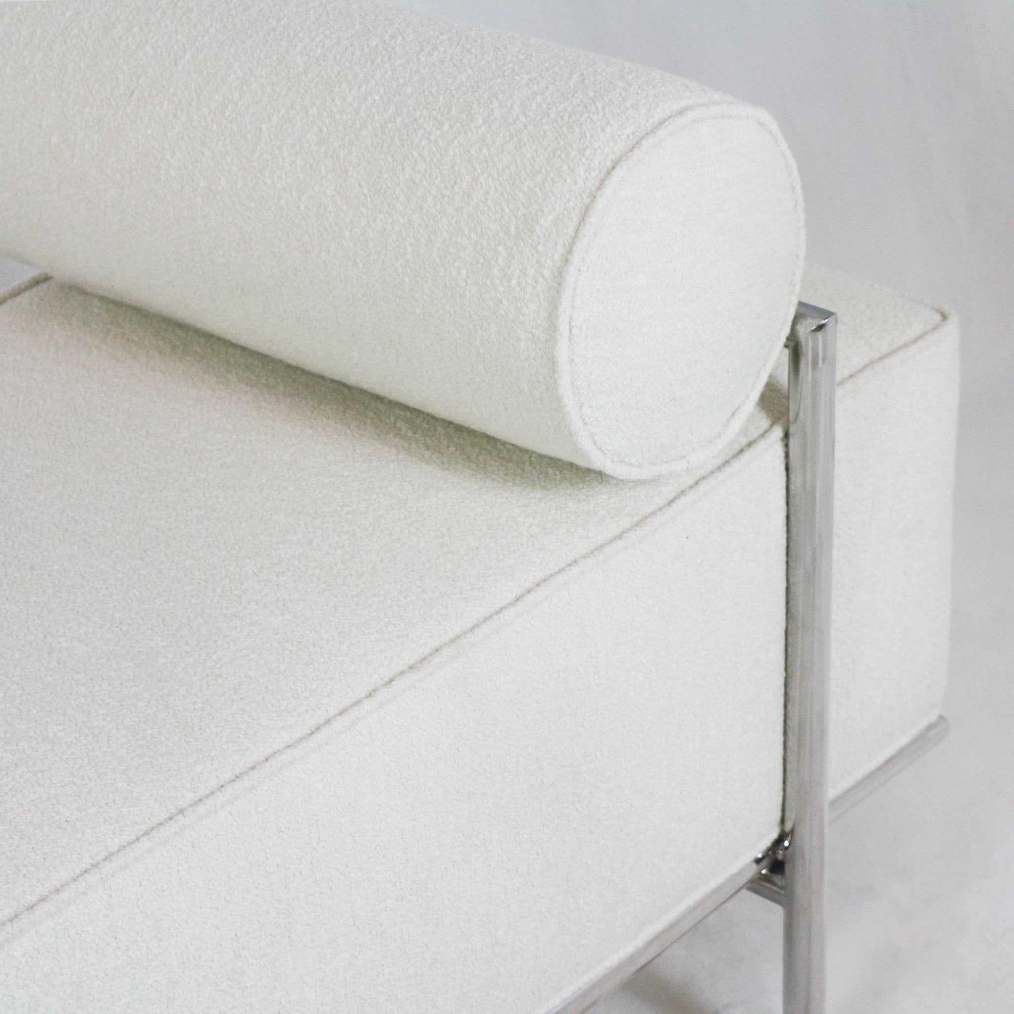 Close-up of an off-white boucle-upholstered bench end with a cylindrical bolster pillow, visible stitched seams and a polished stainless-steel frame and leg supporting the cushioned seat.