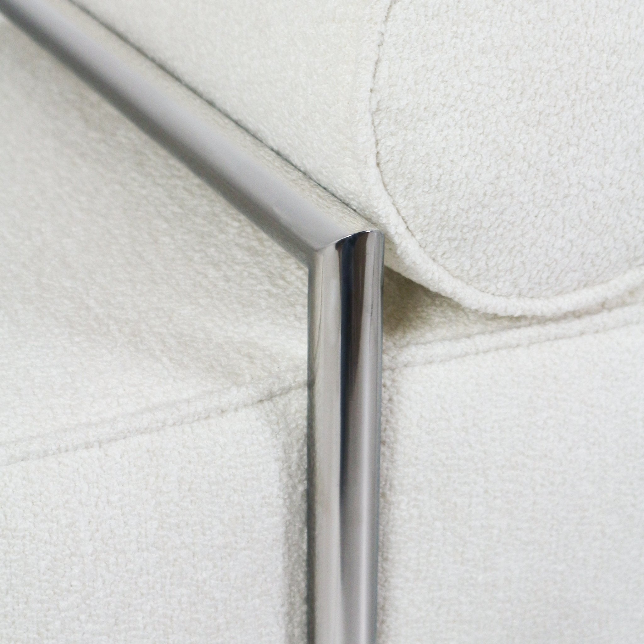 Close-up of a polished chrome metal leg and frame against off-white boucle fabric upholstery, showing the rounded bolster cushion and textured stitched seam.