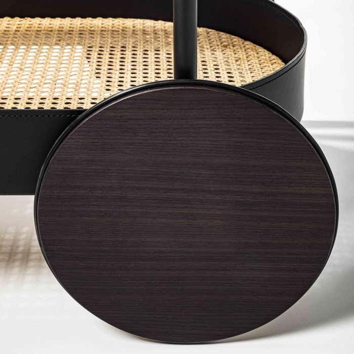 Close-up of a bar cart showing a large dark wood wheel, matte black metal frame and a woven natural rattan shelf edge.