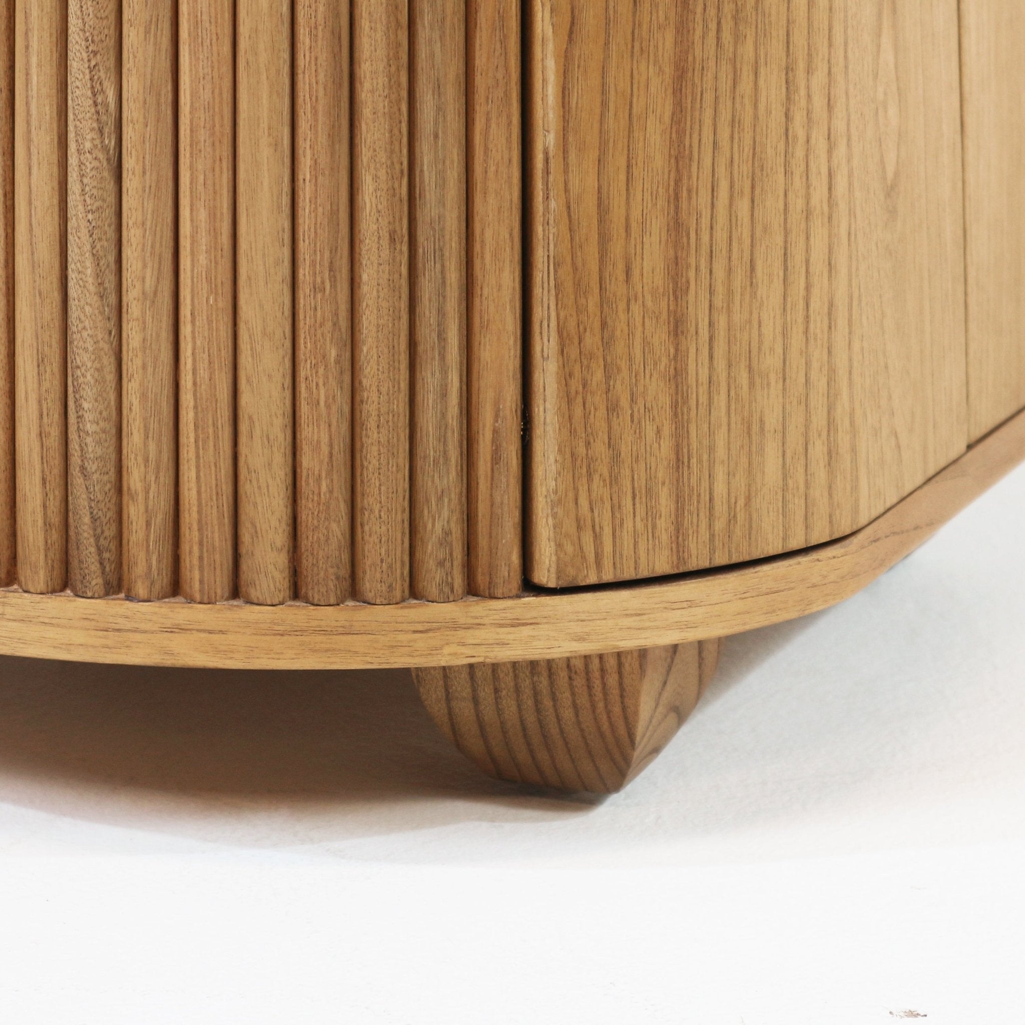 Close-up of a natural solid wood nightstand showing vertical fluted slats, a curved corner trim and a small tapered wooden foot in a light natural finish.