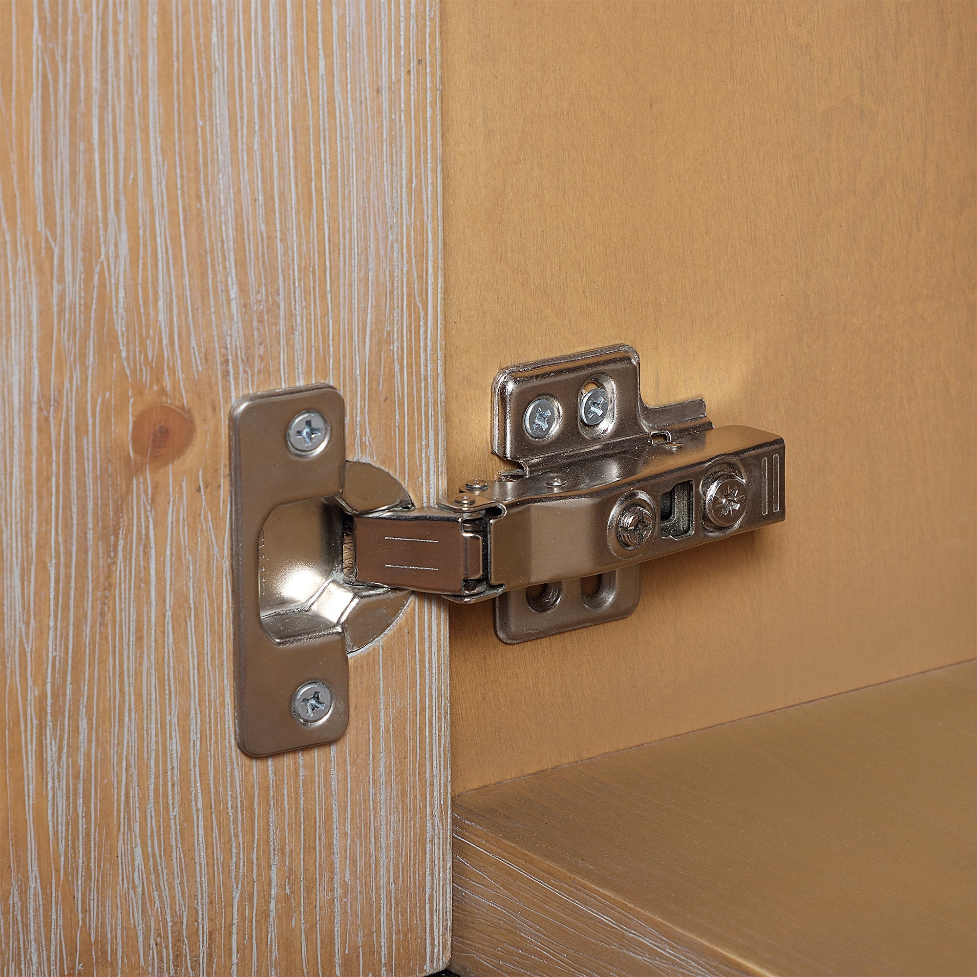 Close-up of a metal soft-close cabinet hinge mounted to light brown natural wood vanity door and interior shelf, showing the mounting plates, screws, and wood grain texture.