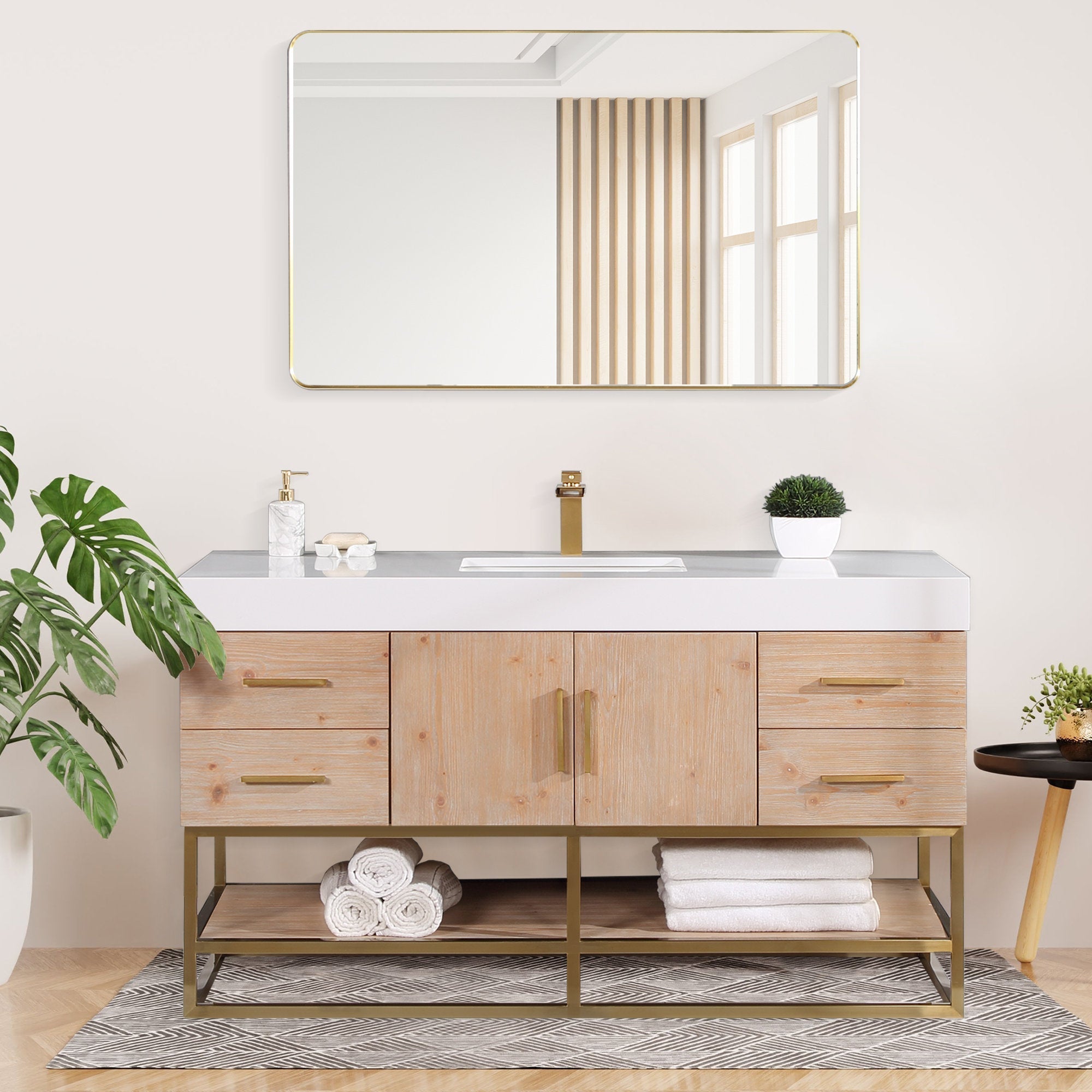 Modern single-sink bathroom vanity in light natural wood with a white composite stone countertop, rectangular undermount sink, brass faucet and hardware, lower open shelf with folded towels, gold-framed mirror and potted plants.