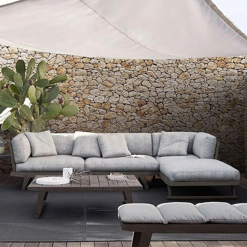 Outdoor teak-framed sectional sofa with light gray fabric cushions and chaise, paired with a low teak coffee table and cushioned bench against a stone wall and shade sail in a coastal-contemporary style.