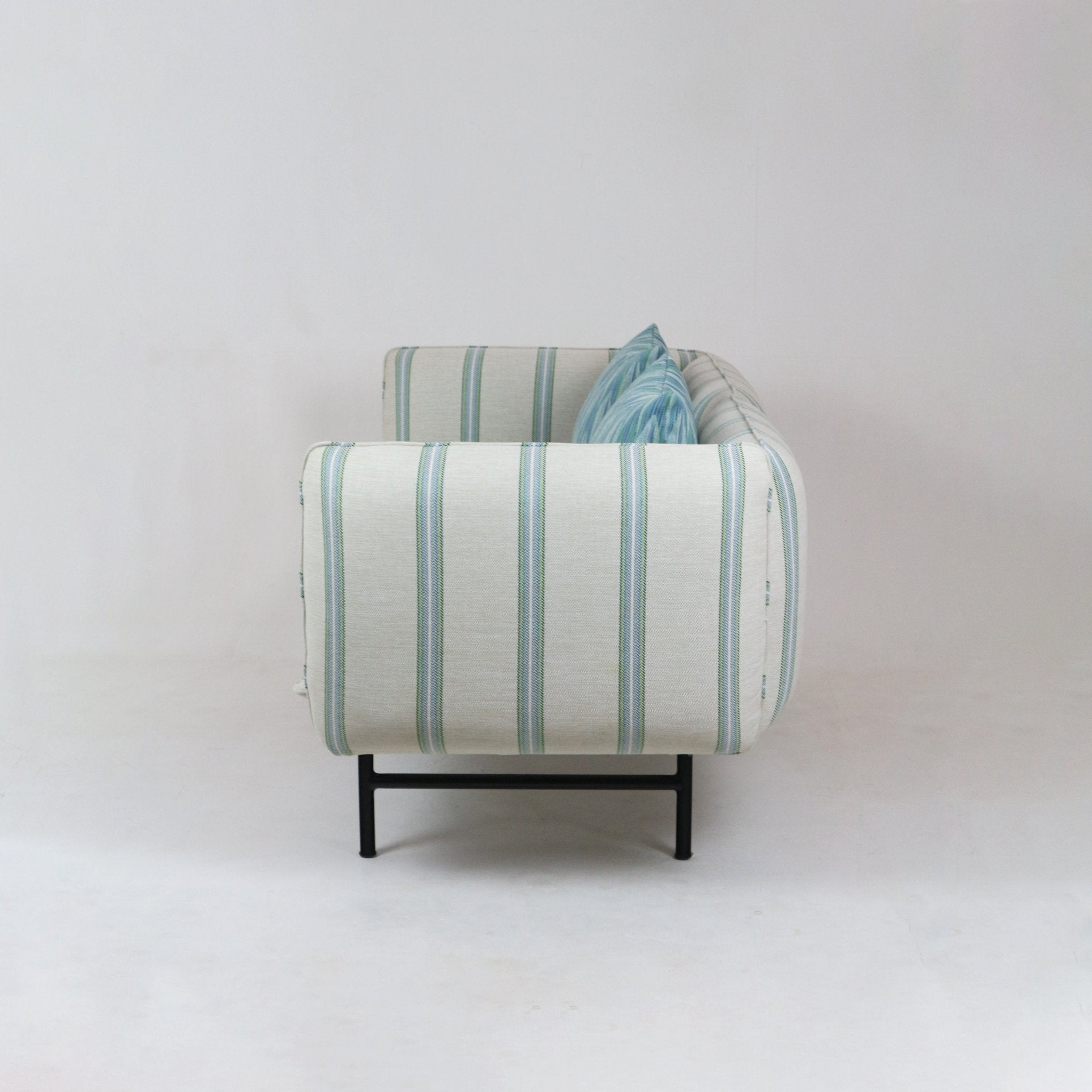Side view of an outdoor sectional sofa upholstered in cream fabric with blue-green vertical stripes, a blue patterned throw pillow visible, and black metal legs in a modern coastal style.