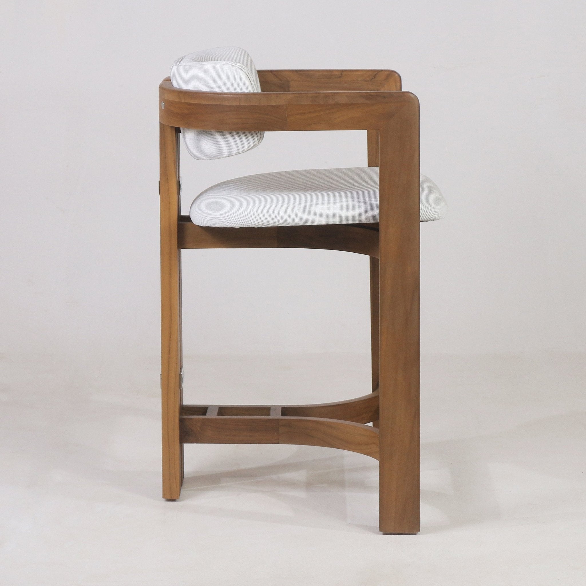 Side profile of a teak outdoor bar stool showing warm teak wood frame with curved arm and built-in footrest, white upholstered seat and small cushioned back against a neutral background.