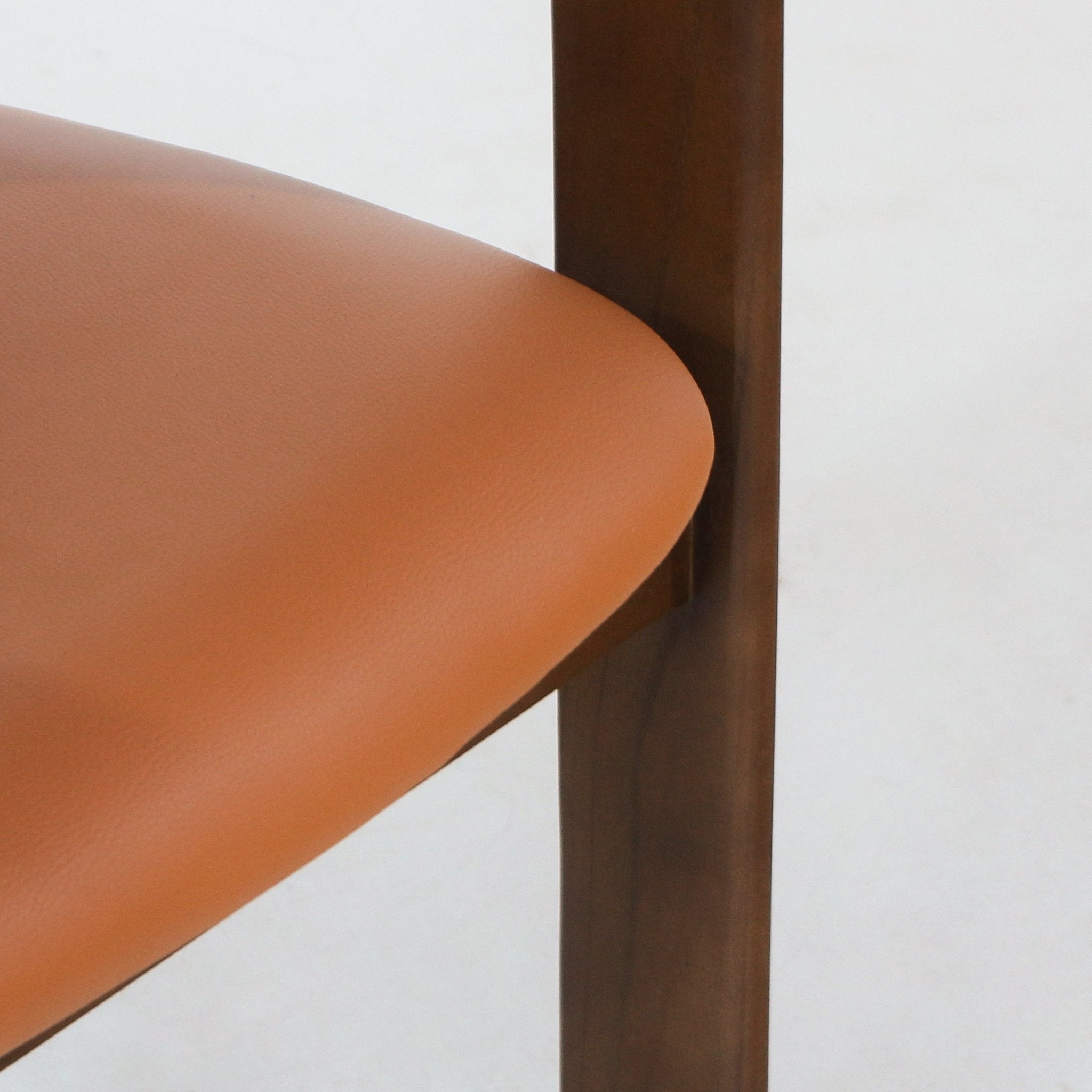 Close-up of a tan leather dining chair seat and curved edge, highlighting smooth leather upholstery and a walnut wood leg and vertical support in a Mid-Century Modern style against a neutral background.