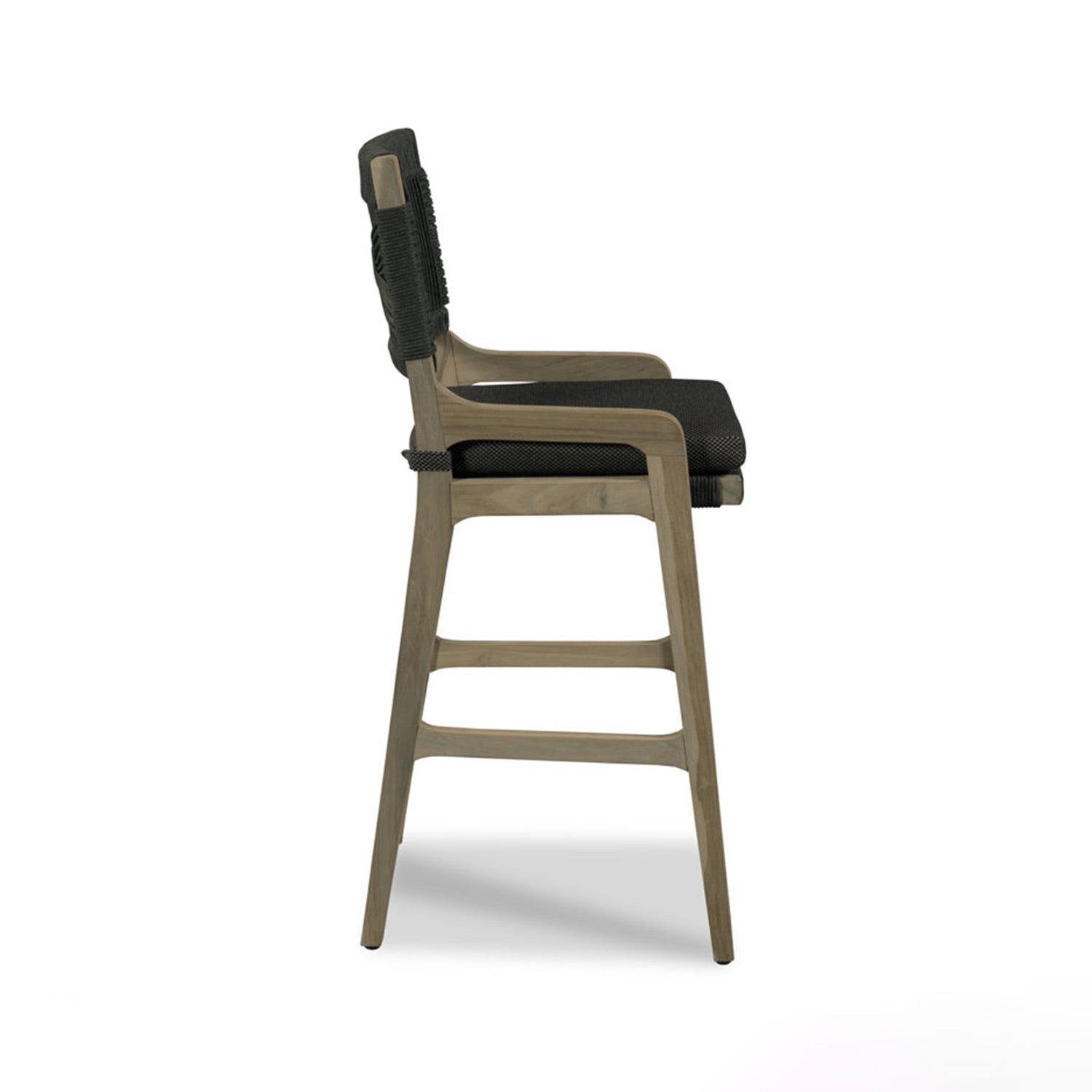 Side profile of a natural teak outdoor bar stool with tapered legs, built-in footrests and curved armrests, featuring a black fabric seat cushion and a woven black rope back in a coastal-contemporary style.