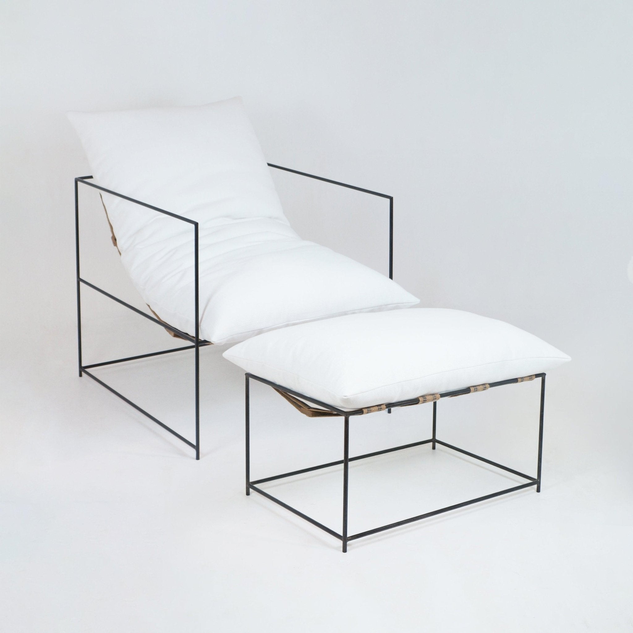 Modern lounge chair and ottoman with thin black metal frame, plush white linen cushions and natural leather suspension straps, photographed on a neutral white background.