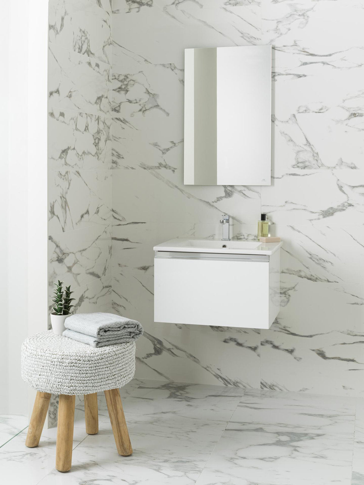 Modern 24.5-inch single-sink floating vanity with stone top and white solid-wood cabinet mounted on a marble-tiled wall, rectangular mirror above; knitted stool with wooden legs and folded towels beside it.