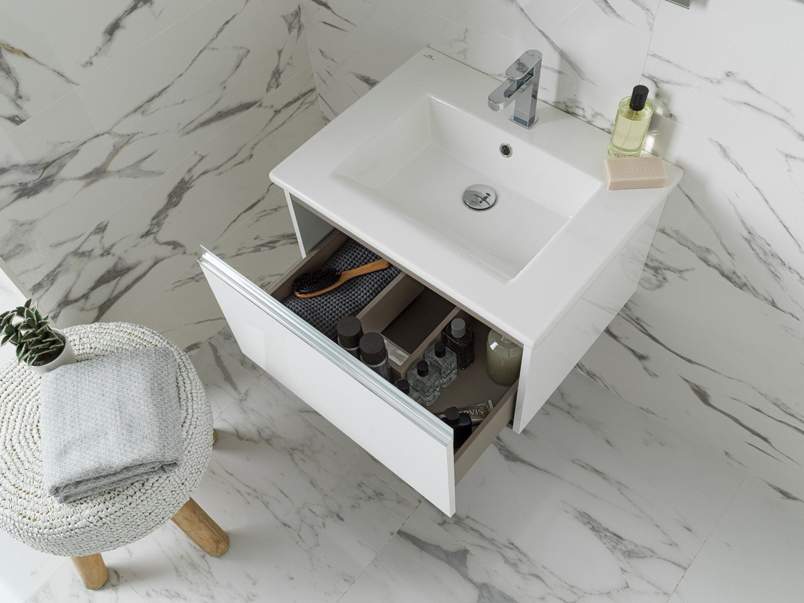 Top-down view of a modern solid wood single-sink vanity with a stone top and white integrated basin, chrome faucet and open drawer showing organized bottles and a hairbrush; marble-look tile floor and a woven stool with towel and plant beside it.