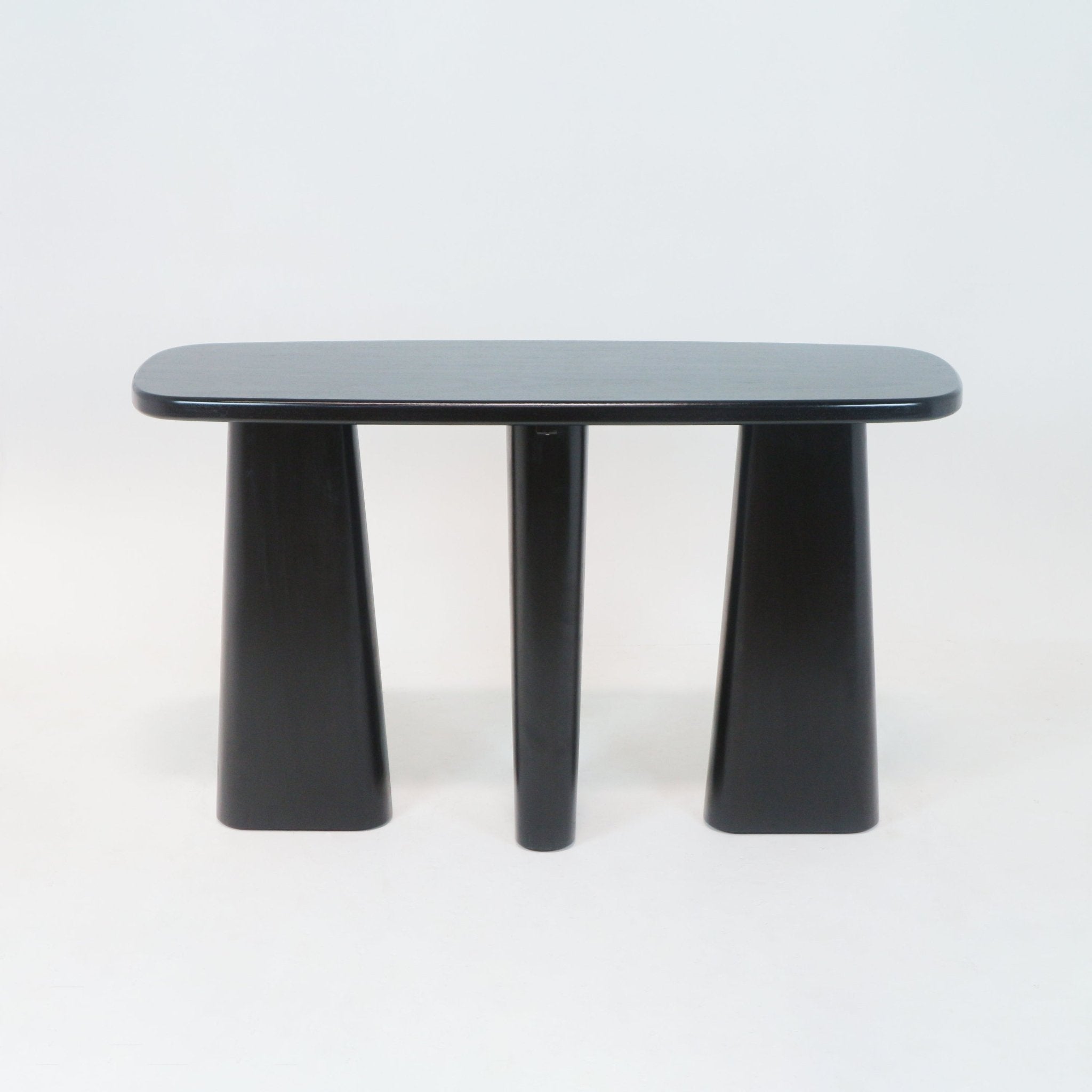 Minimalist black solid wood console table with a rounded rectangular top and three pedestal legs — two flared block bases and a central cylindrical leg, shown on a white background.