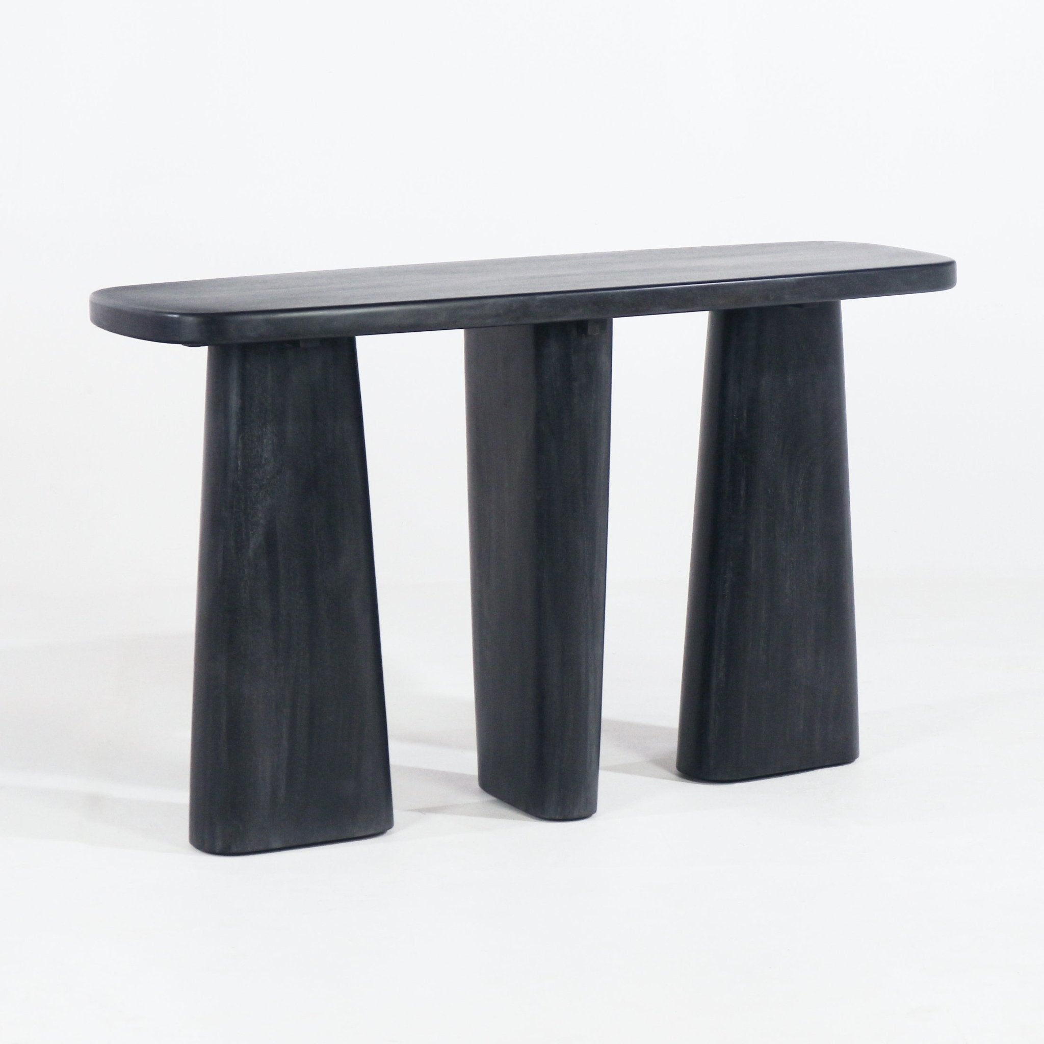 Smoked-black solid wood console table with a rounded rectangular top and three thick tapered slab legs, showcasing a modern minimalist silhouette against a white background.