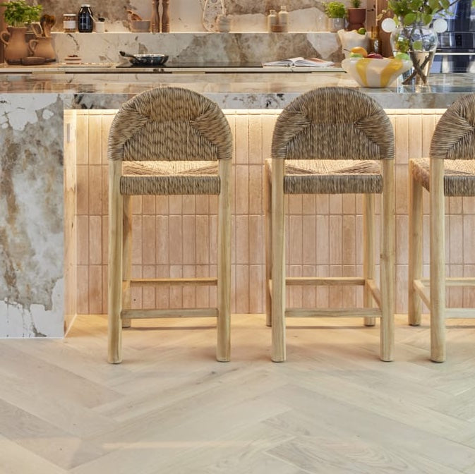 Three oak counter stools with woven natural rope seats and rounded backs sit at a marble-topped kitchen island with warm under-counter lighting and herringbone wood floor.