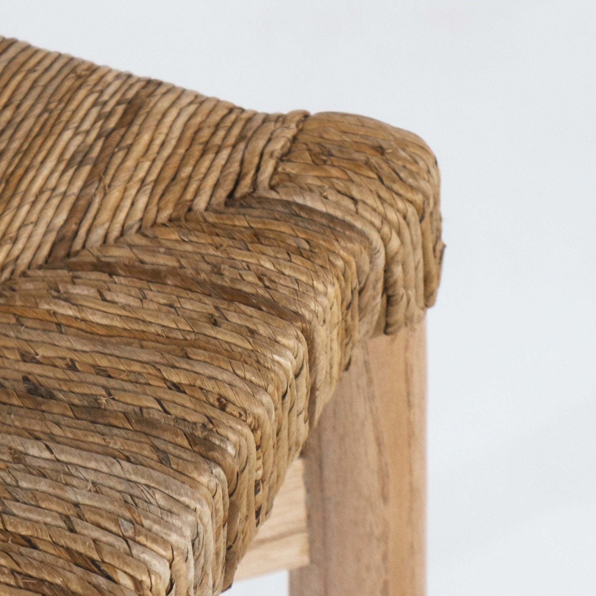 Close-up of a natural seagrass woven seat wrapped over the corner of a teak counter stool, highlighting the braided texture and warm natural teak leg.
