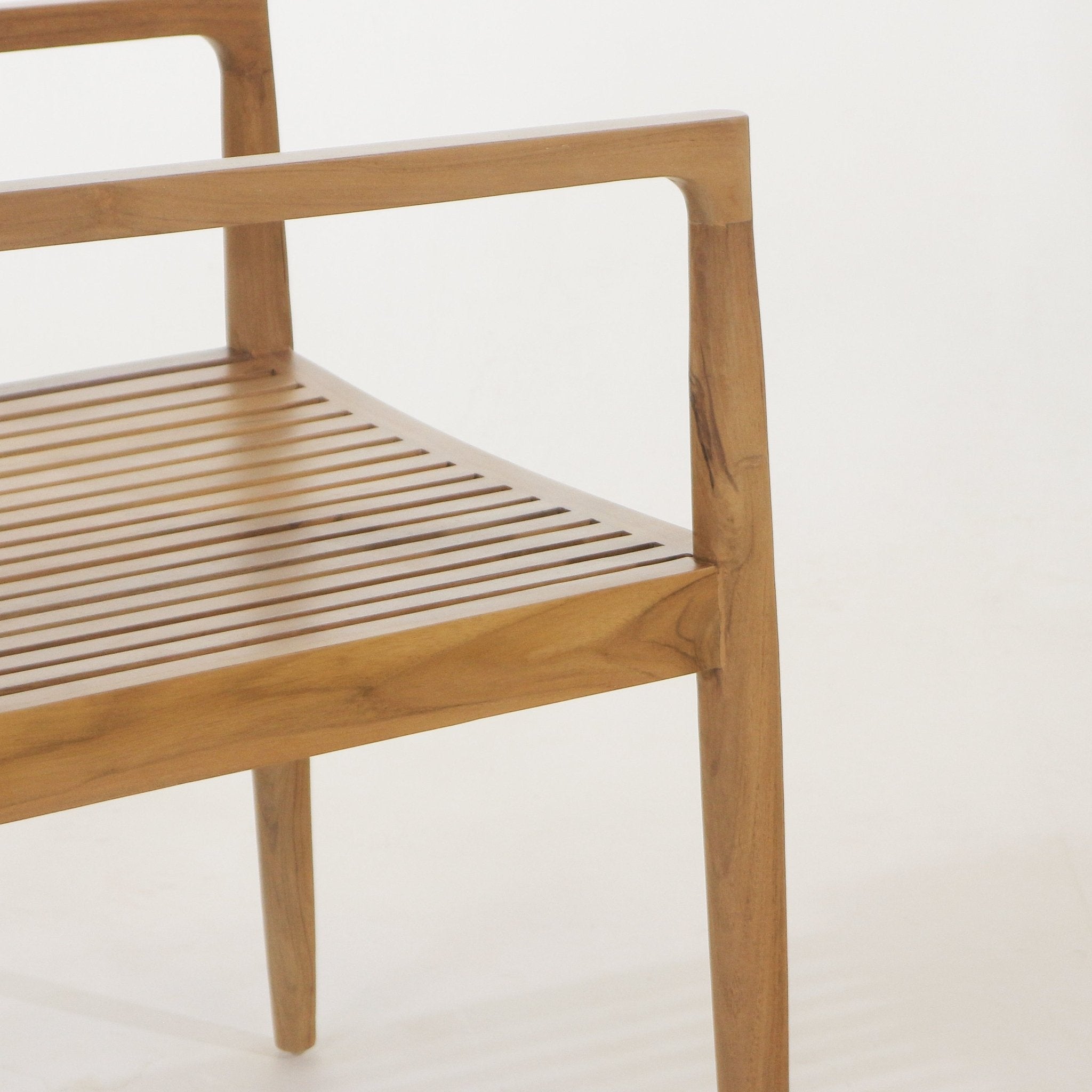 Close-up of a natural teak outdoor dining chair showing the slatted seat, angled armrest and tapered leg with warm brown wood grain and smooth sculpted joinery.