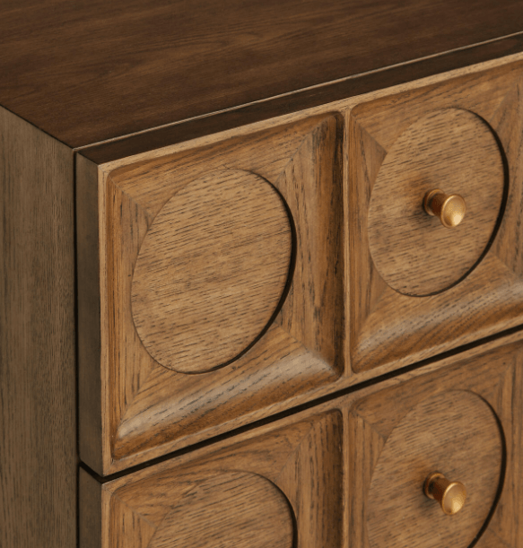 Close-up of a solid wood nightstand showing carved drawer fronts with recessed circular panels and round brass knobs in a warm brown finish, highlighting the contemporary glam detailing.