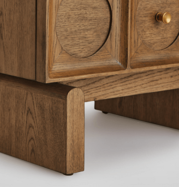 Close-up of the lower corner of a solid wood nightstand showing the rounded pedestal base, carved circular drawer-front detail and a small brass knob in a warm natural wood finish.