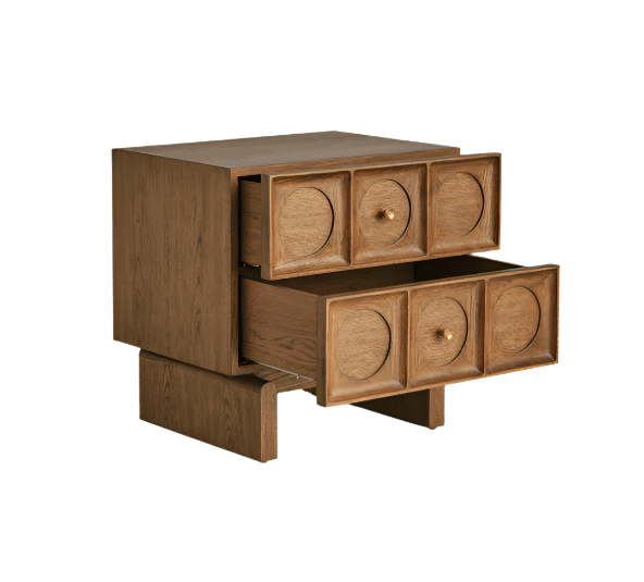 Solid wood nightstand in warm brown natural finish with two stacked drawers featuring carved circular recessed panels and small brass knobs, lower drawer pulled open to show interior, contemporary glam bedside table.