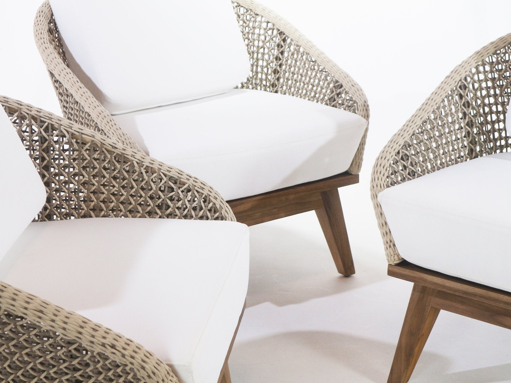 Close-up of three coastal-style outdoor accent chairs featuring beige rope-woven arms, white UV-resistant fabric cushions, and angled teak wood bases.