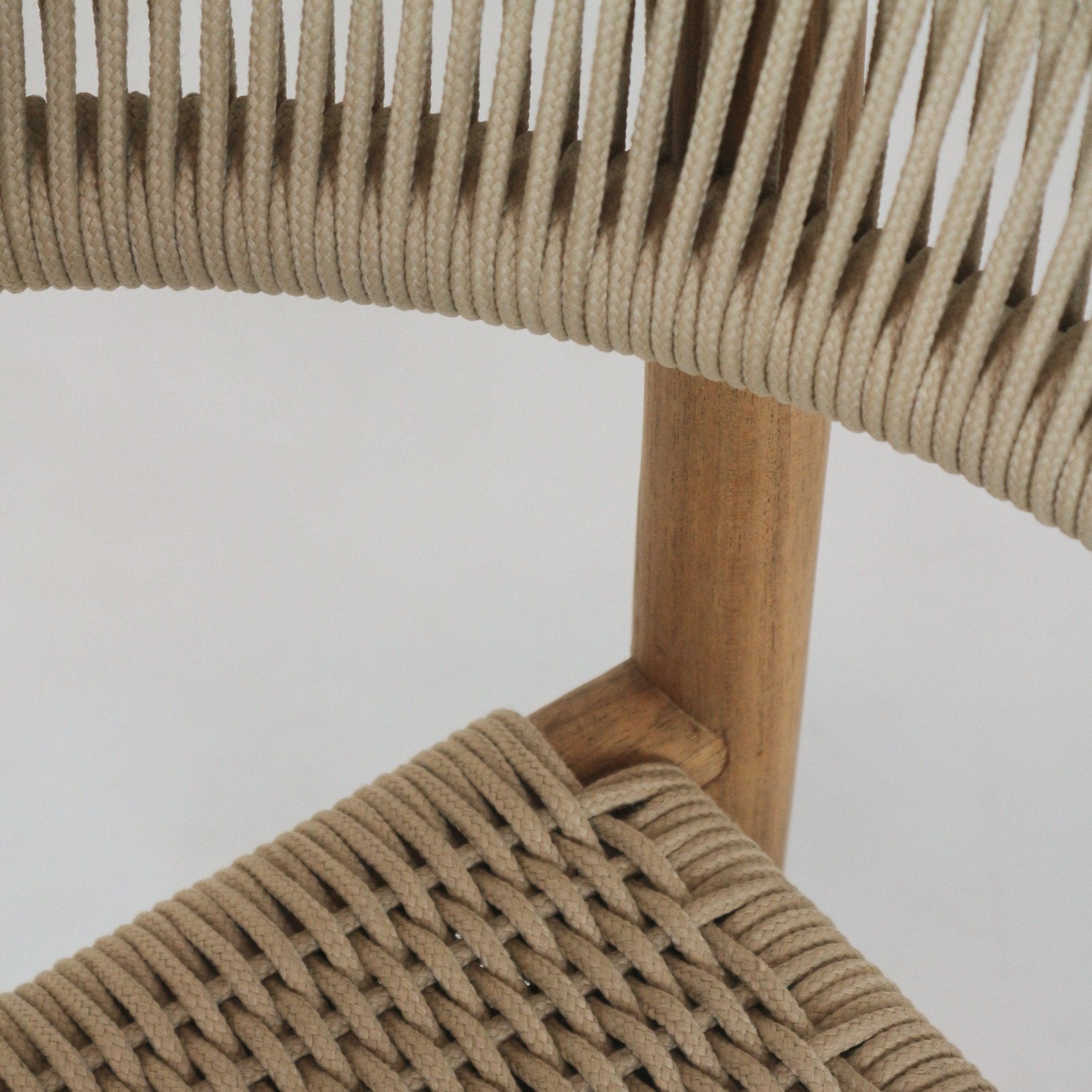 Close-up of a coastal-style teak dining chair showing beige polypropylene rope woven seat and back with a rounded teak leg and visible joint detail.