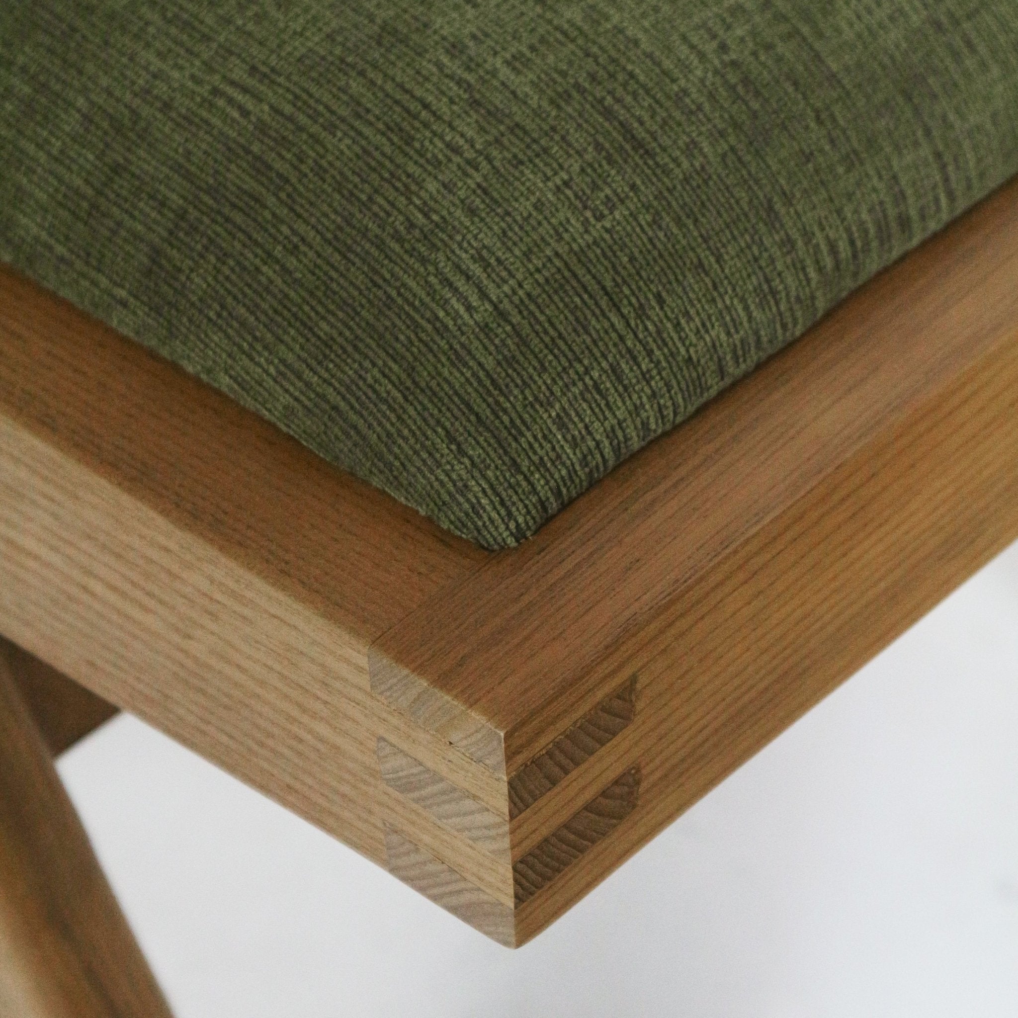 Close-up of a green chenille upholstered seat cushion on a natural ash wood frame, highlighting the corner finger joint and warm wood grain.