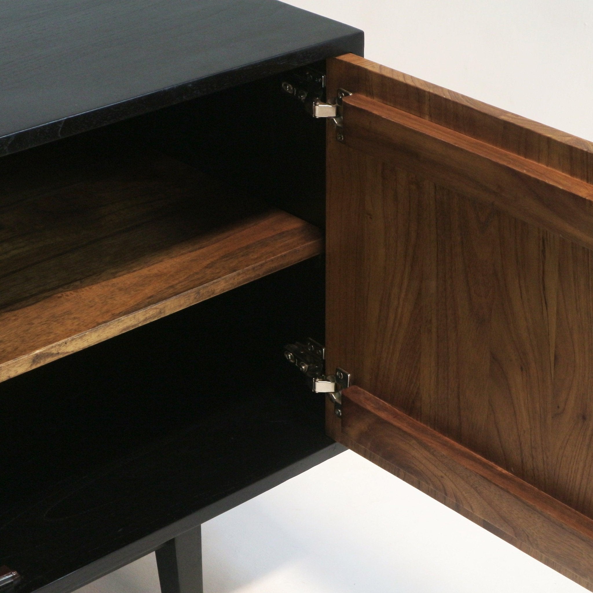 Close-up of a teak sideboard with an open door revealing a solid wood teak shelf and exposed metal hinges, showing a dark top and black interior in a mid-century modern style.